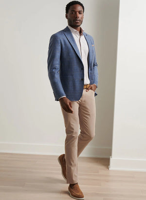 A man stands indoors wearing the Peter Millar Delrose Plaid Soft Jacket, a white dress shirt, beige trousers, and brown shoes on a light wood floor.