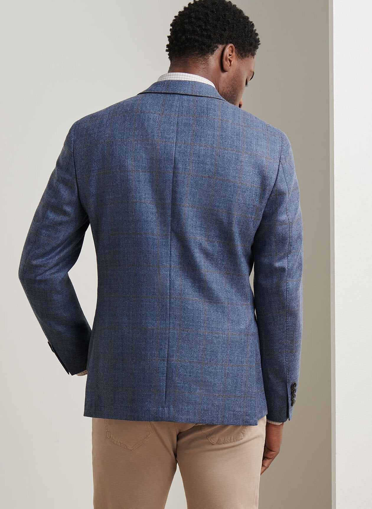 A man wearing the Peter Millar Delrose Plaid Soft Jacket and beige pants stands facing away from the camera against a light-colored wall.