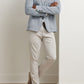 A person stands with arms crossed, wearing the Peter Millar Barclay Knit Soft Jacket in light gray over a white shirt, paired with beige pants and beige loafers, leaning against a white wall on a light wood floor.