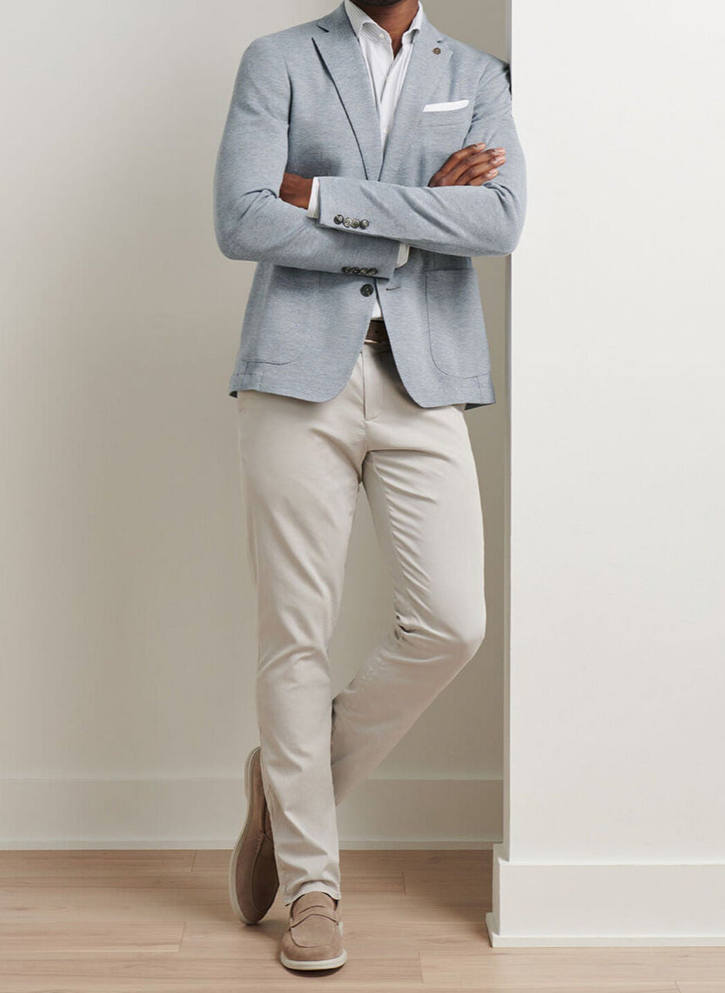 A person stands with arms crossed, wearing the Peter Millar Barclay Knit Soft Jacket in light gray over a white shirt, paired with beige pants and beige loafers, leaning against a white wall on a light wood floor.