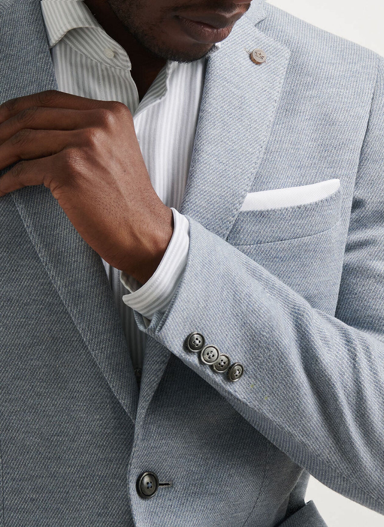 A man adjusts the lapel of the Peter Millar Barclay Knit Soft Jacket in light grey, featuring a white pocket square and worn over a white striped dress shirt.