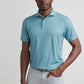 A man stands against a plain background wearing the Peter Millar Corinth Performance Jersey Polo in light blue with four-way stretch and light grey pants, one hand casually in his pocket.