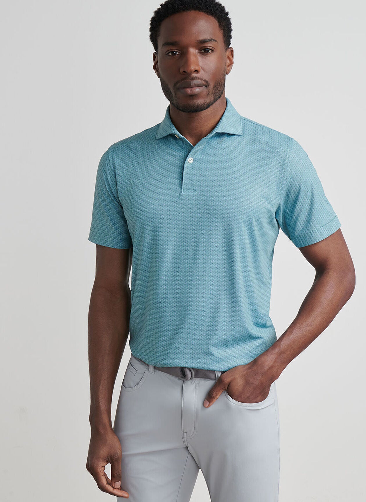 A man stands against a plain background wearing the Peter Millar Corinth Performance Jersey Polo in light blue with four-way stretch and light grey pants, one hand casually in his pocket.