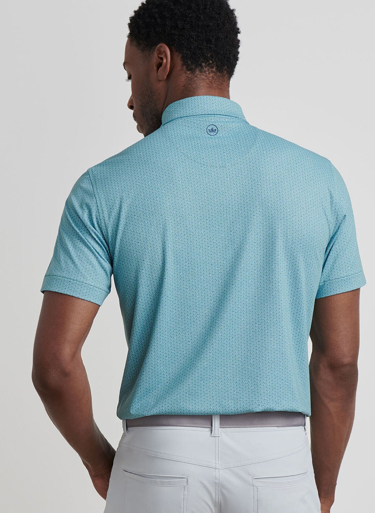 A man faces away, wearing the Peter Millar Corinth Performance Jersey Polo in light blue with UPF 50+ sun protection and light gray pants, set against a plain background.