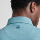 A close-up of a man's head and shoulders highlights the Peter Millar Corinth Performance Jersey Polo in teal pattern, featuring a blue crown logo below the collar and UPF 50+ sun protection.
