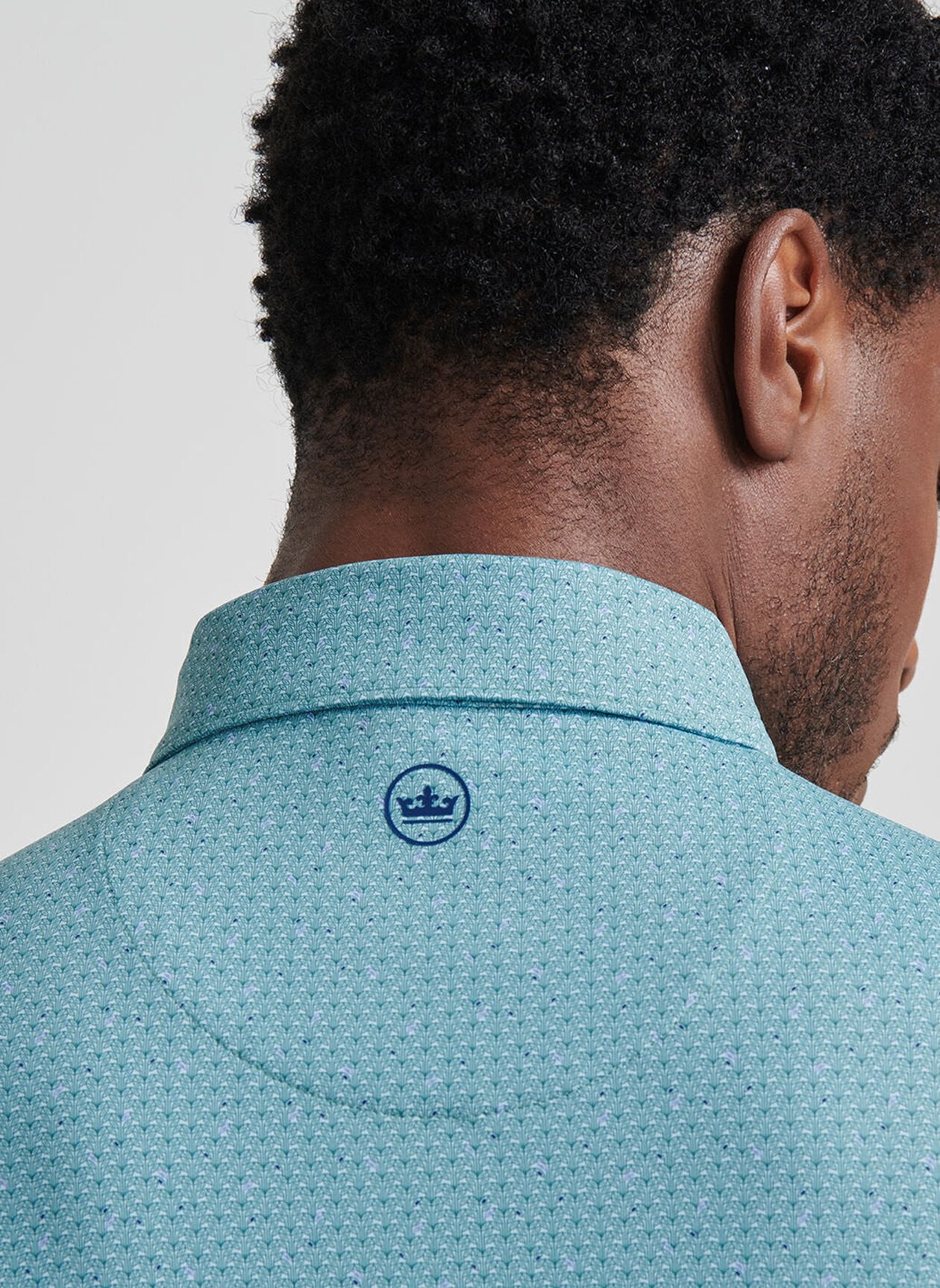 A close-up of a man's head and shoulders highlights the Peter Millar Corinth Performance Jersey Polo in teal pattern, featuring a blue crown logo below the collar and UPF 50+ sun protection.