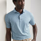 A man wearing the Peter Millar Chase Performance Jersey Polo in light blue stripes and beige pants stands against a white wall, looking to the side with one hand in his pocket.