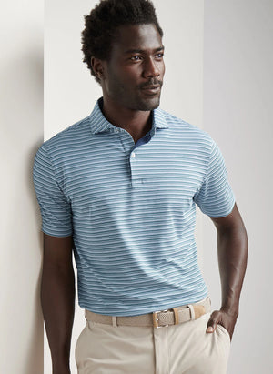 A man wearing the Peter Millar Chase Performance Jersey Polo in light blue stripes and beige pants stands against a white wall, looking to the side with one hand in his pocket.