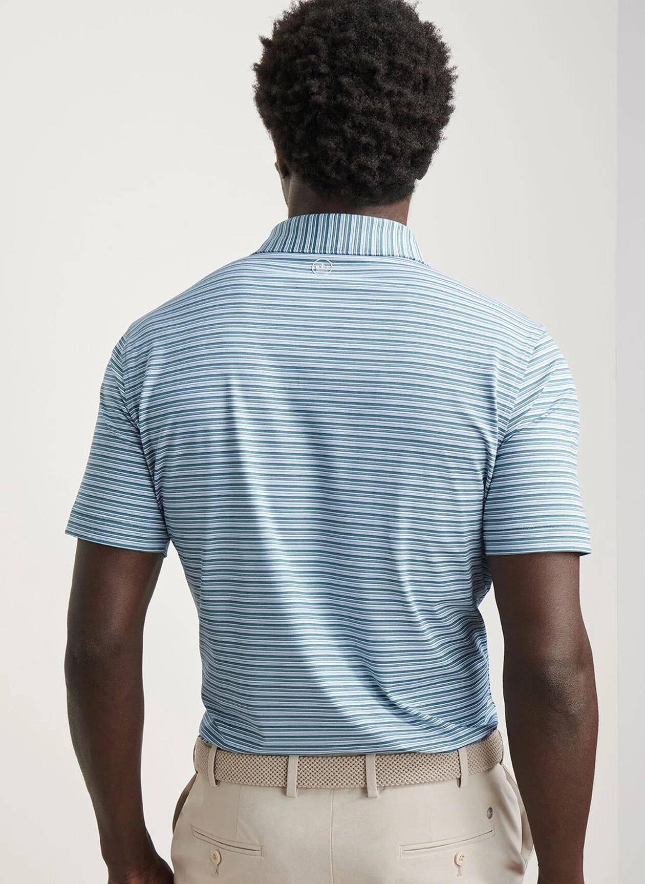 A man, seen from behind against a plain background, wears the Peter Millar Chase Performance Jersey Polo in light blue and navy stripes with four-way stretch, paired with beige pants.