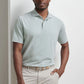 A man stands against a white wall, wearing the Peter Millar Albatross Cotton-Blend Pique Polo in light gray, tucked into beige pants with a beige belt, one hand in his pocket.