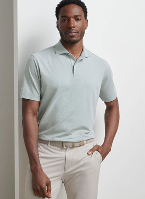 A man stands against a white wall, wearing the Peter Millar Albatross Cotton-Blend Pique Polo in light gray, tucked into beige pants with a beige belt, one hand in his pocket.