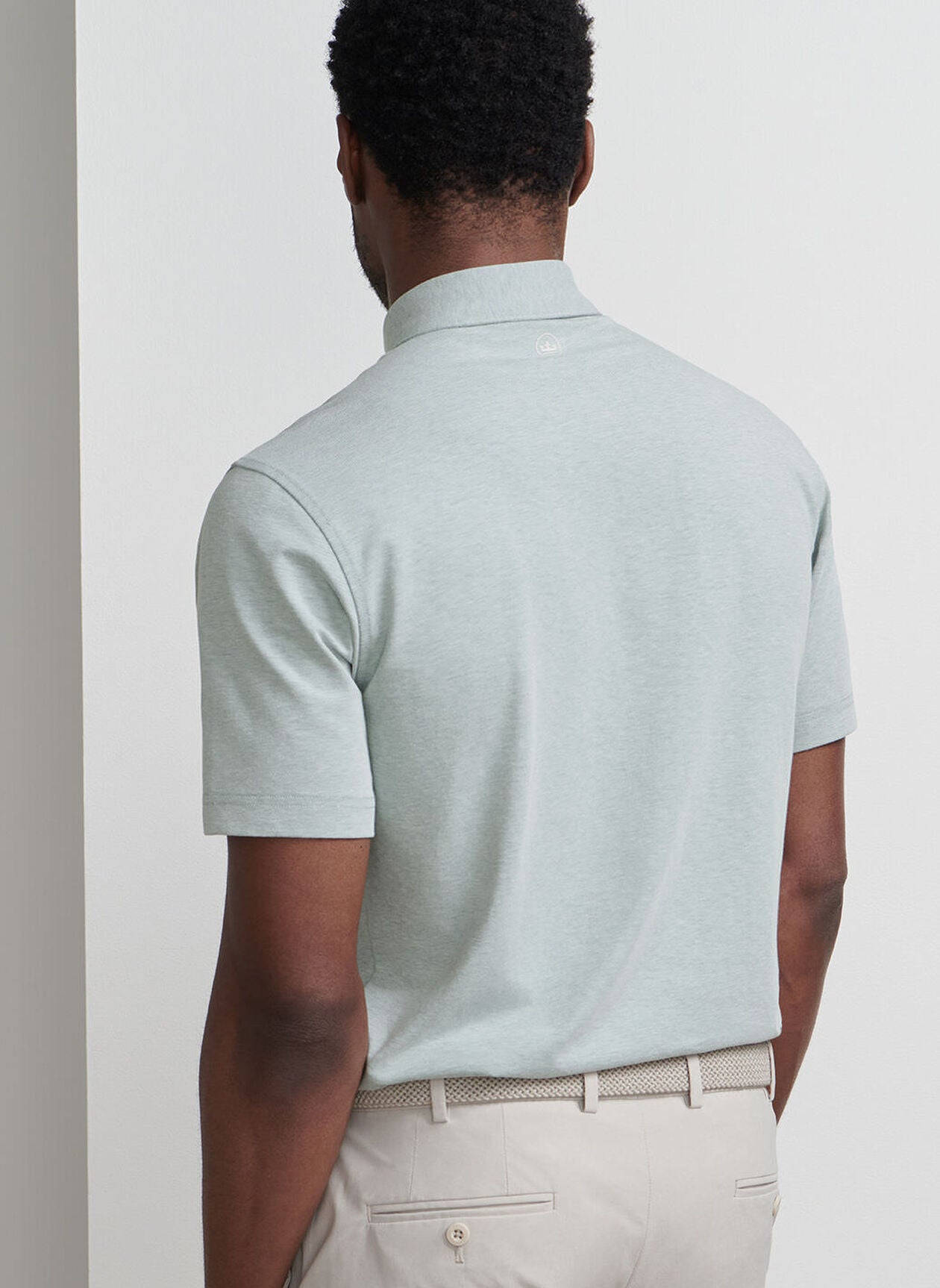 A man faces away from the camera, wearing a light gray Peter Millar Albatross Cotton-Blend Pique Polo, beige pants, and a textured beige belt against a plain light background.
