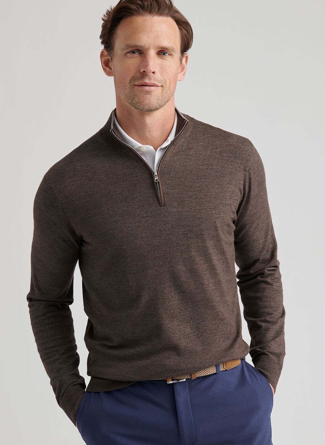 Man wearing a brown Peter Millar Excursionist Flex Quarter-Zip Sweater over a white collared shirt and blue pants, standing with hands in pockets against a plain background.