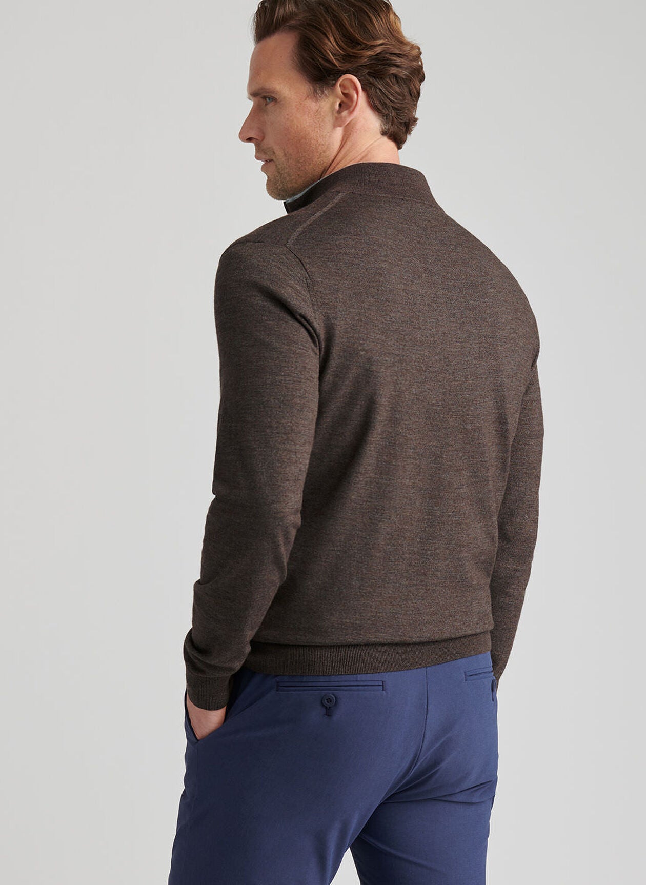 A man faces away, wearing a brown Peter Millar Excursionist Flex Quarter-Zip Sweater and blue pants against a plain light gray background.