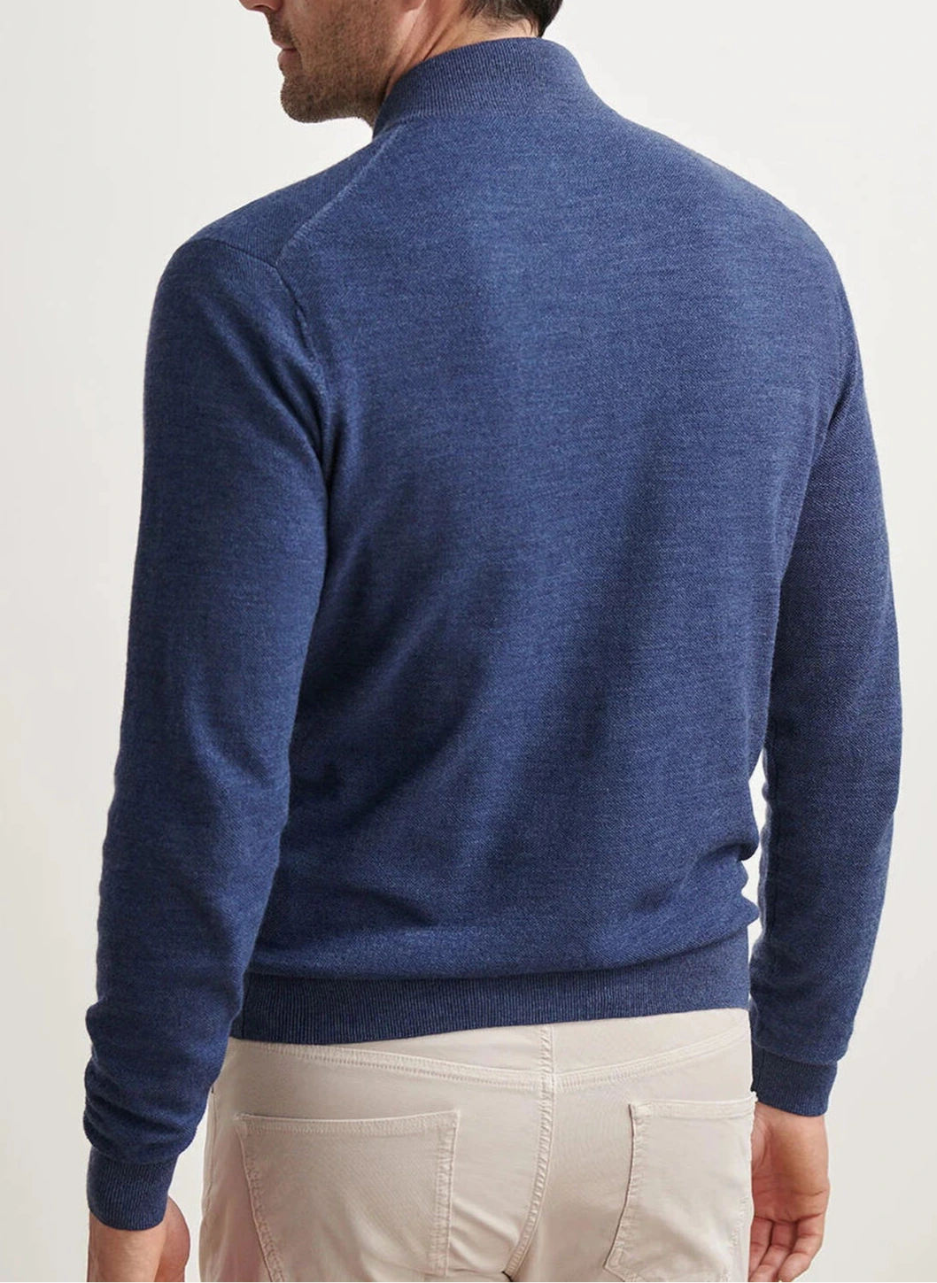 A man wearing a Peter Millar Excursionist Flex Full-Zip Sweater in blue and light beige pants is shown from the back, facing slightly left against a plain background.