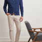 A man wearing a blue Peter Millar Excursionist Flex Full-Zip Sweater, beige pants, and brown slip-on shoes stands next to a gray cushioned wooden chair.