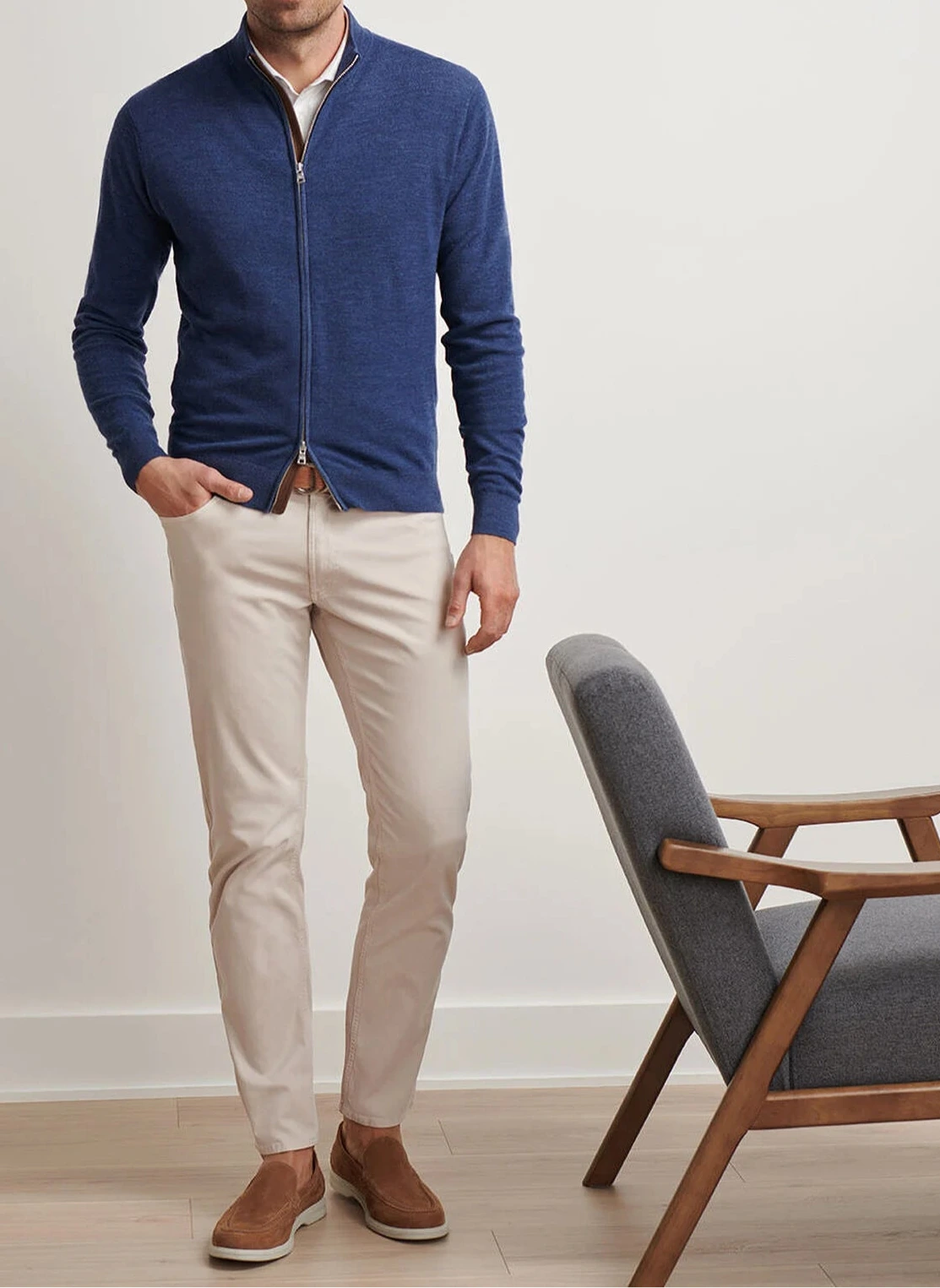 A man wearing a blue Peter Millar Excursionist Flex Full-Zip Sweater, beige pants, and brown slip-on shoes stands next to a gray cushioned wooden chair.