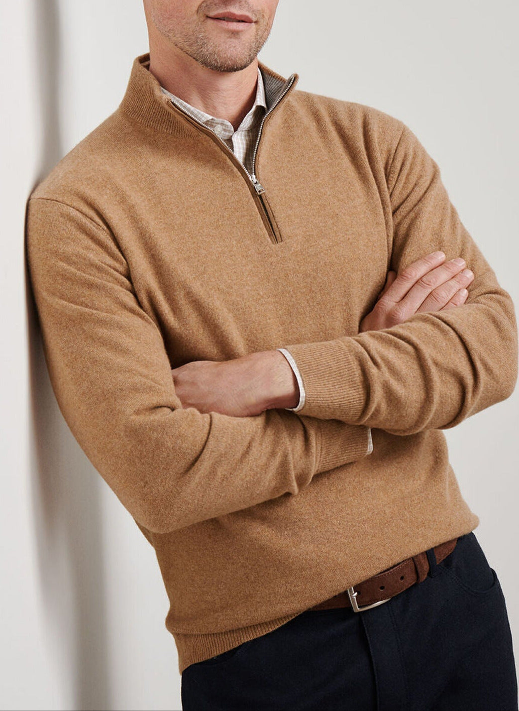 A man stands with his arms crossed over a collared shirt, wearing the Peter Millar Artisan Crafted Cashmere Flex Quarter-Zip in tan, inspired by classic British Isles knit styles.