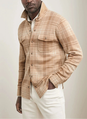 A man in the Peter Millar Gordon Plaid Sweater Shirt Jacket—beige and tan plaid Merino wool with chest pockets—pairs it with white pants for a minimal, versatile style, standing with one hand in his pocket.