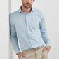 A man in a Peter Millar Bechet Performance Poplin Sport Shirt in light blue check and beige pants stands against a white wall with his hands in his pockets.