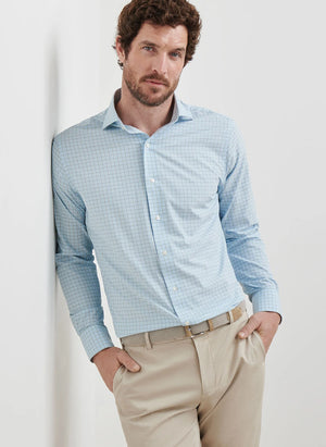 A man in a Peter Millar Bechet Performance Poplin Sport Shirt in light blue check and beige pants stands against a white wall with his hands in his pockets.