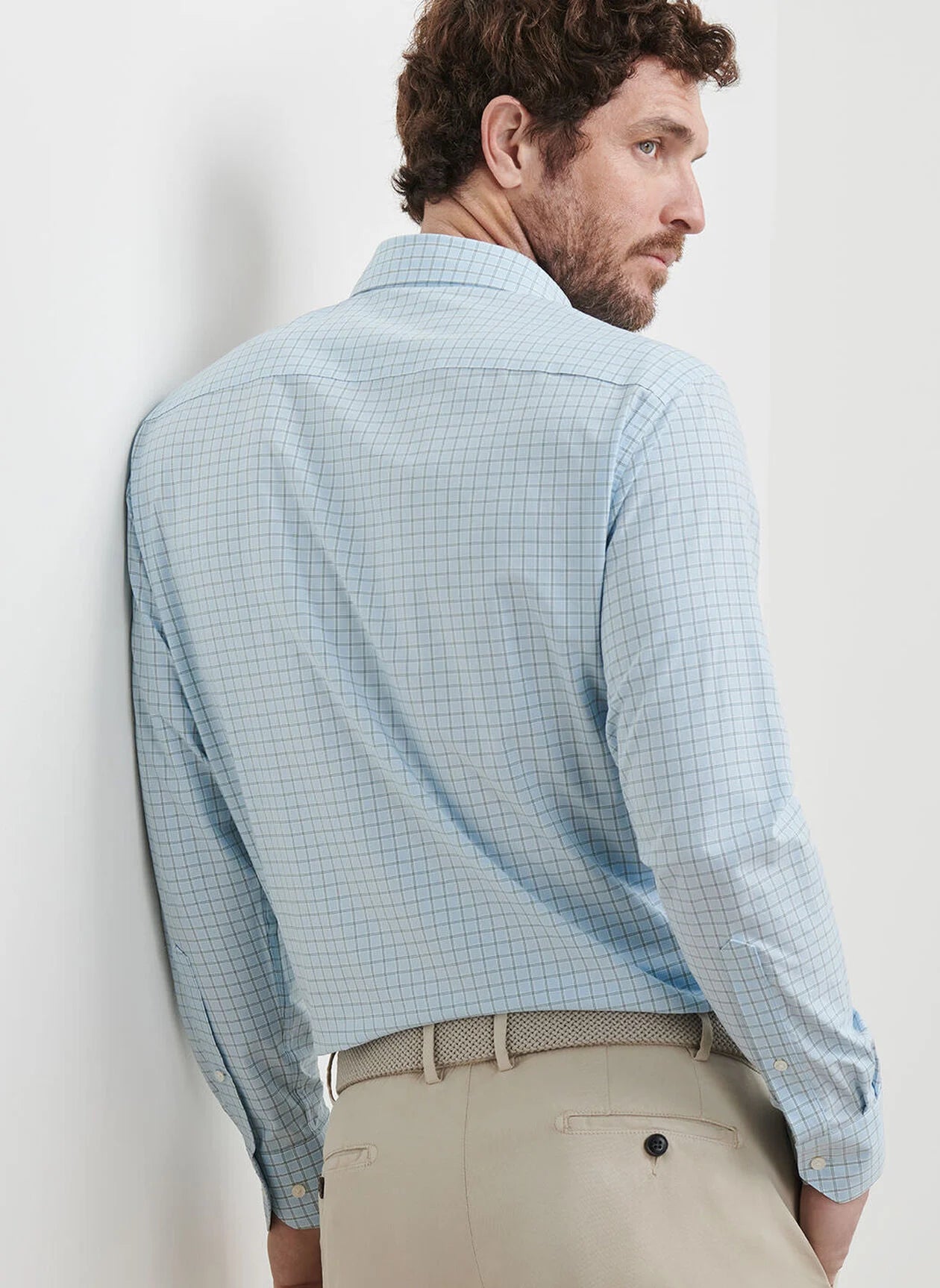 A man stands with his back to the camera, looking over his shoulder, wearing the Peter Millar Bechet Performance Poplin Sport Shirt in light blue checks and beige pants.
