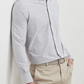 A man wears the Peter Millar Carter Performance Poplin Sport Shirt in light gray plaid, paired with beige dress pants and a woven belt, standing with one hand in his pocket against a white background.