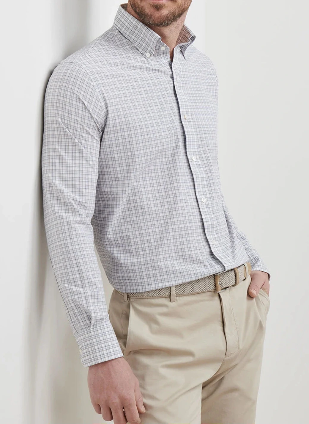A man wears the Peter Millar Carter Performance Poplin Sport Shirt in light gray plaid, paired with beige dress pants and a woven belt, standing with one hand in his pocket against a white background.