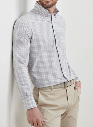 A man wears the Peter Millar Carter Performance Poplin Sport Shirt in light gray plaid, paired with beige dress pants and a woven belt, standing with one hand in his pocket against a white background.