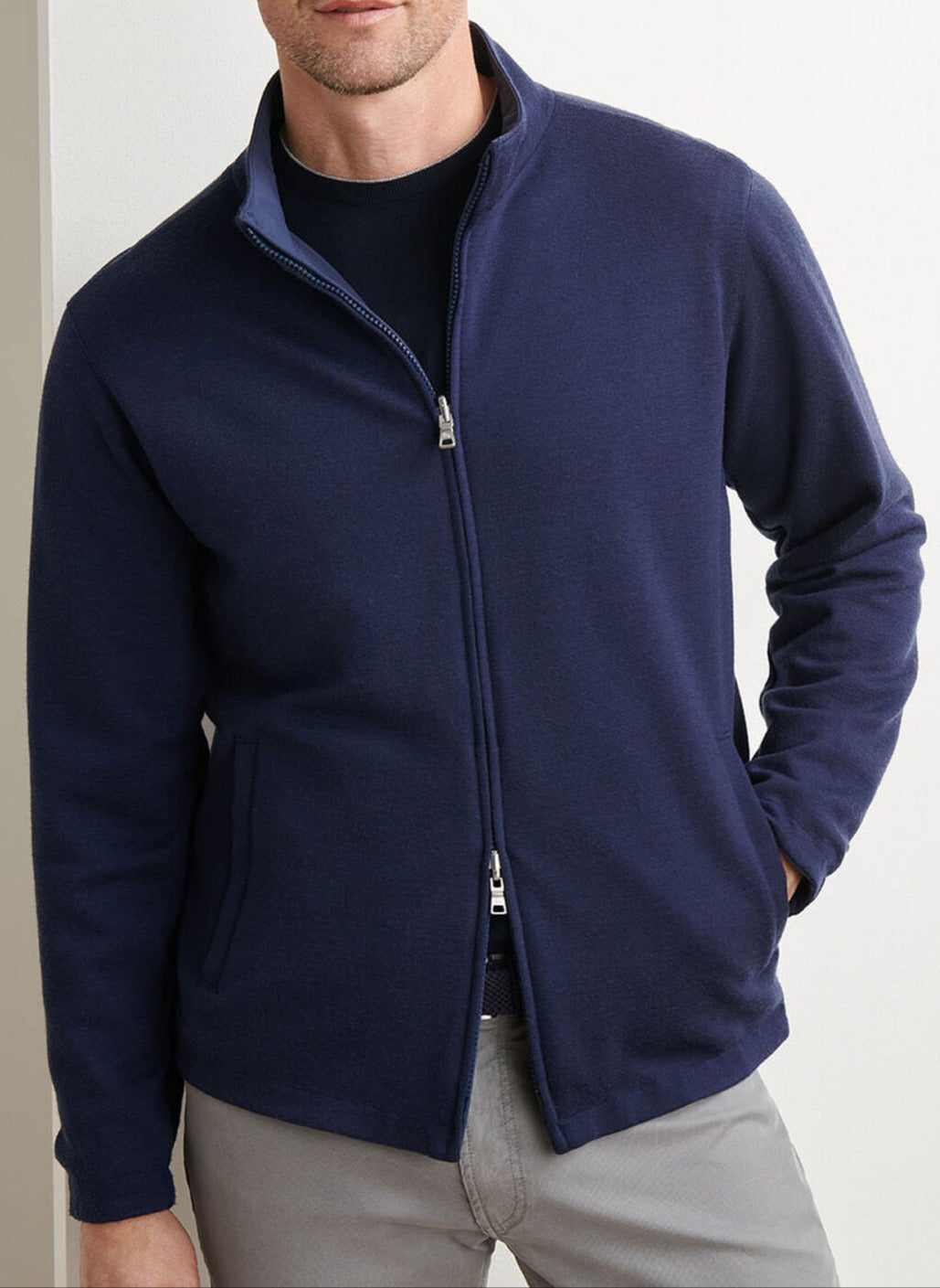 A man wears the Peter Millar Ryder Reversible Jacket in navy over a black shirt and light gray pants, standing with one hand in his pocket.