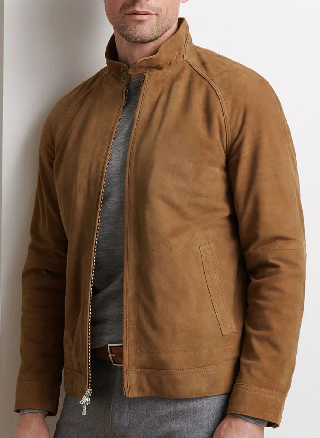 A man wears the Peter Millar Soho Suede Bomber, a lightweight jacket with a microfiber lining, over a grey shirt against a plain light-colored background.