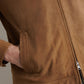 Close-up of a person wearing the Soho Suede Bomber by Peter Millar in tan, featuring lightweight suede, a zipper, and a hand in the front pocket. The background is plain and neutral.