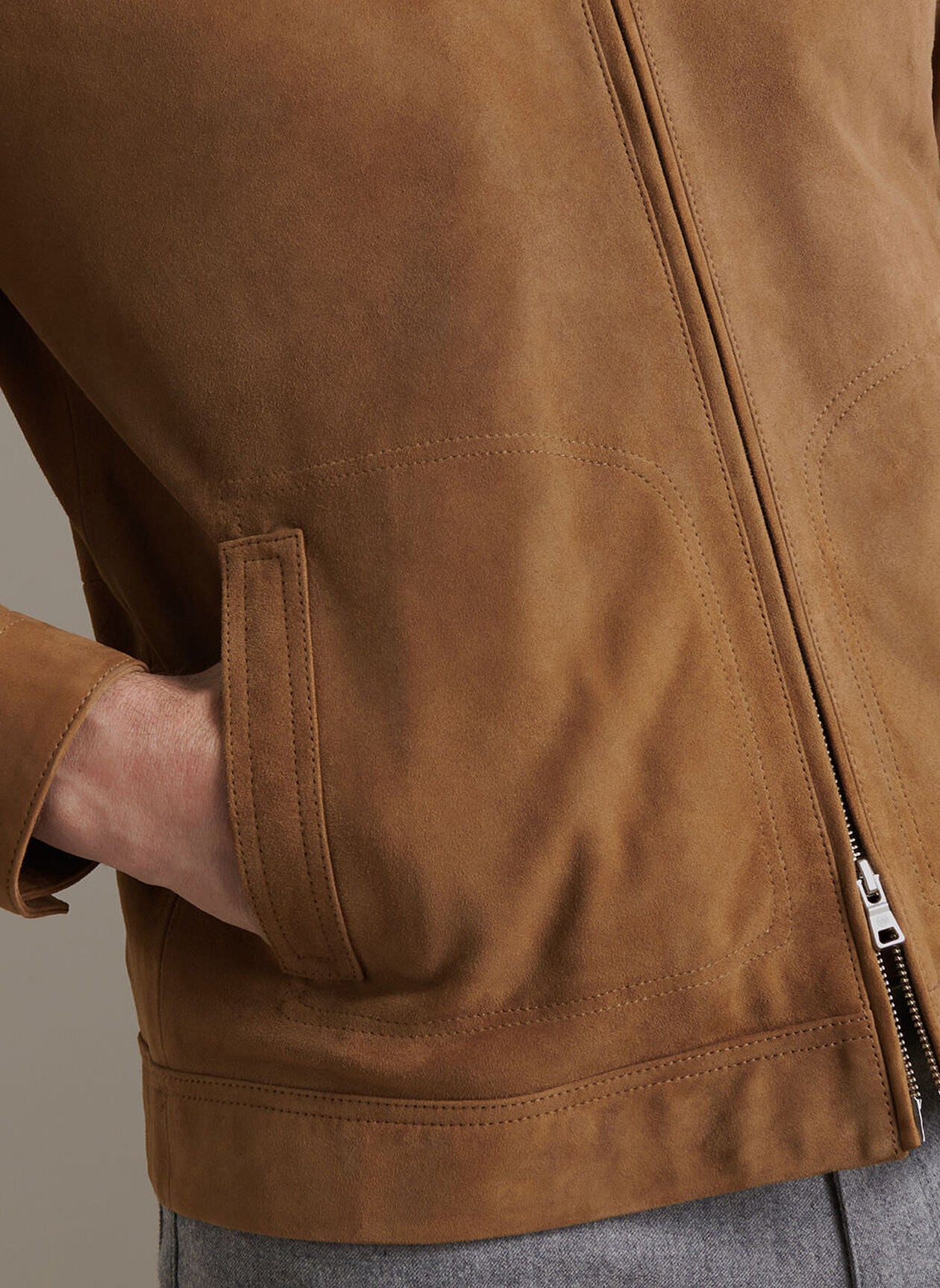Close-up of a person wearing the Soho Suede Bomber by Peter Millar in tan, featuring lightweight suede, a zipper, and a hand in the front pocket. The background is plain and neutral.