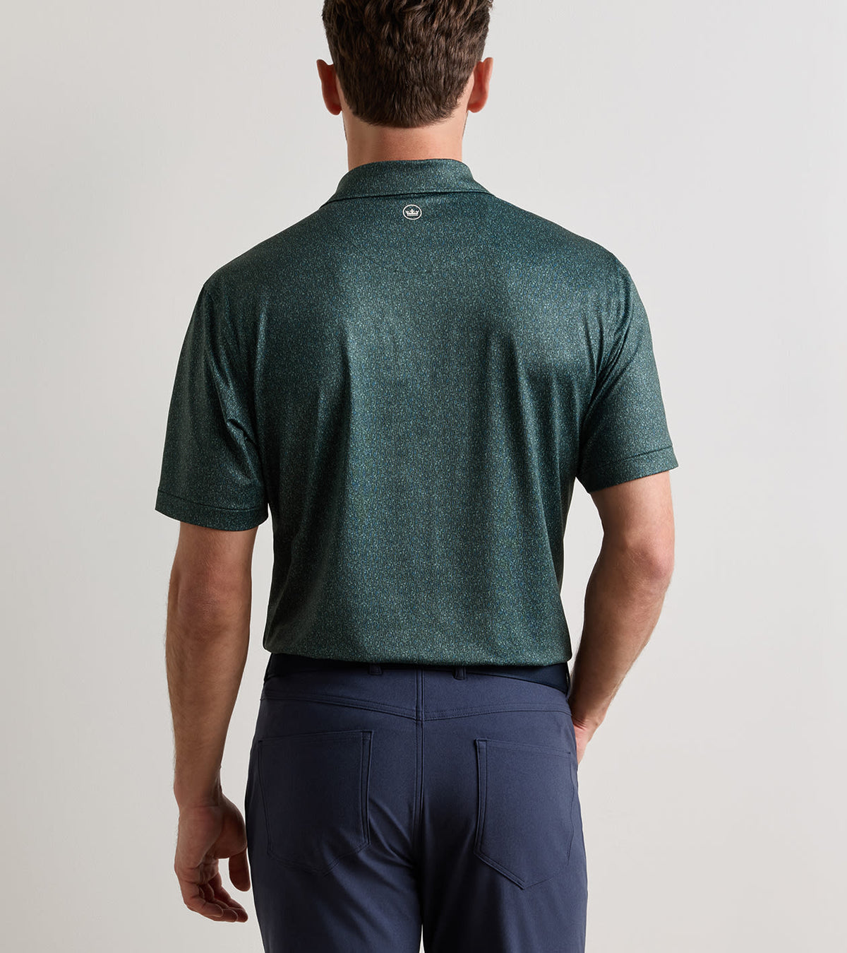 Back view of a person wearing a dark green, four-way stretch patterned Transfusion Performance Jersey Polo and navy blue trousers.
