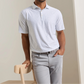 A man wearing the Peter Millar On The Line Performance Jersey Polo—a light gray, short-sleeve collared shirt with a subtle pattern and four-way stretch—paired with gray pants, leans against the back of a light wooden chair.