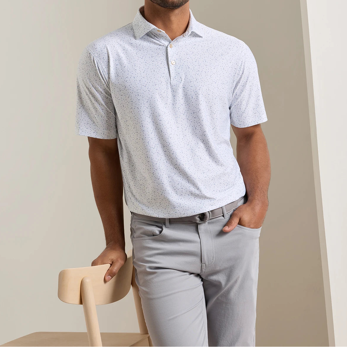 A man wearing the Peter Millar On The Line Performance Jersey Polo—a light gray, short-sleeve collared shirt with a subtle pattern and four-way stretch—paired with gray pants, leans against the back of a light wooden chair.