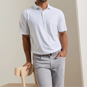A man wearing the Peter Millar On The Line Performance Jersey Polo—a light gray, short-sleeve collared shirt with a subtle pattern and four-way stretch—paired with gray pants, leans against the back of a light wooden chair.