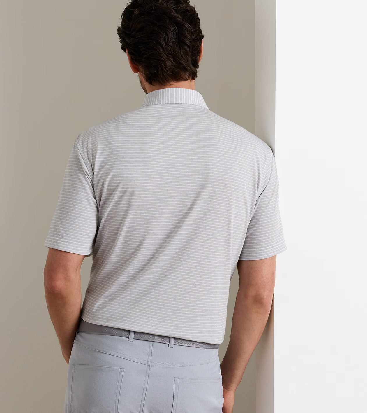 A man stands with his back to the camera, wearing the Peter Millar ETC Stripe Performance Jersey Polo in light gray along with matching pants against a neutral background, embodying effortless Summer Comfort.