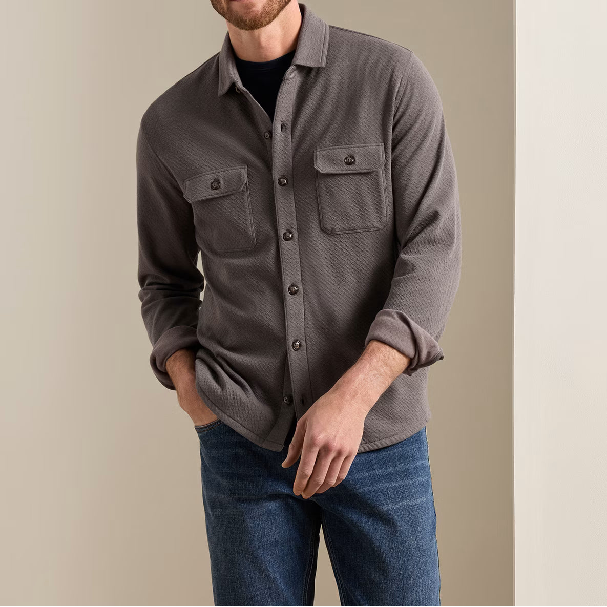 A man wearing a grey Peter Millar Ridgeline Knit Shirt Jacket with two chest pockets over a black t-shirt and blue jeans.