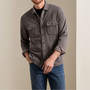 A man wearing a grey Peter Millar Ridgeline Knit Shirt Jacket with two chest pockets over a black t-shirt and blue jeans.