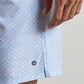 A close-up of a person’s hand resting near the Peter Millar Shoreline Rays Swim Trunk, featuring a blue patterned design with a small crown logo above the hem, highlights the elastic drawstring waistband and quick-drying fabric for enhanced comfort and style.