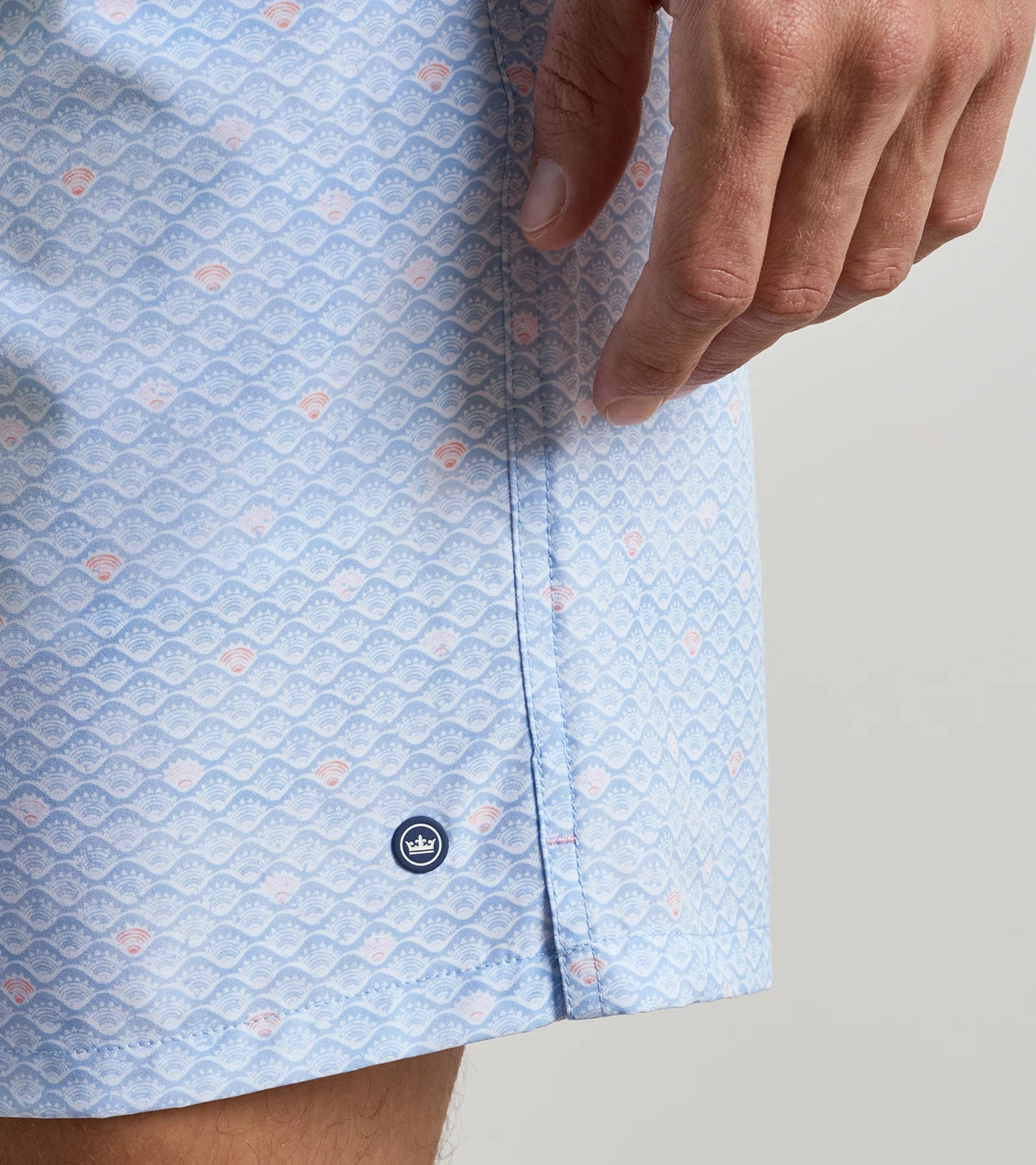 A close-up of a person’s hand resting near the Peter Millar Shoreline Rays Swim Trunk, featuring a blue patterned design with a small crown logo above the hem, highlights the elastic drawstring waistband and quick-drying fabric for enhanced comfort and style.