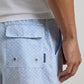 Wearing the Peter Millar Shoreline Rays Swim Trunk in a light blue patterned design, this person sports shorts with a back pocket, metal grommets, and a small navy label. Crafted from quick-drying fabric with an elastic drawstring waistband for comfort, the look is completed with a partially visible white shirt.