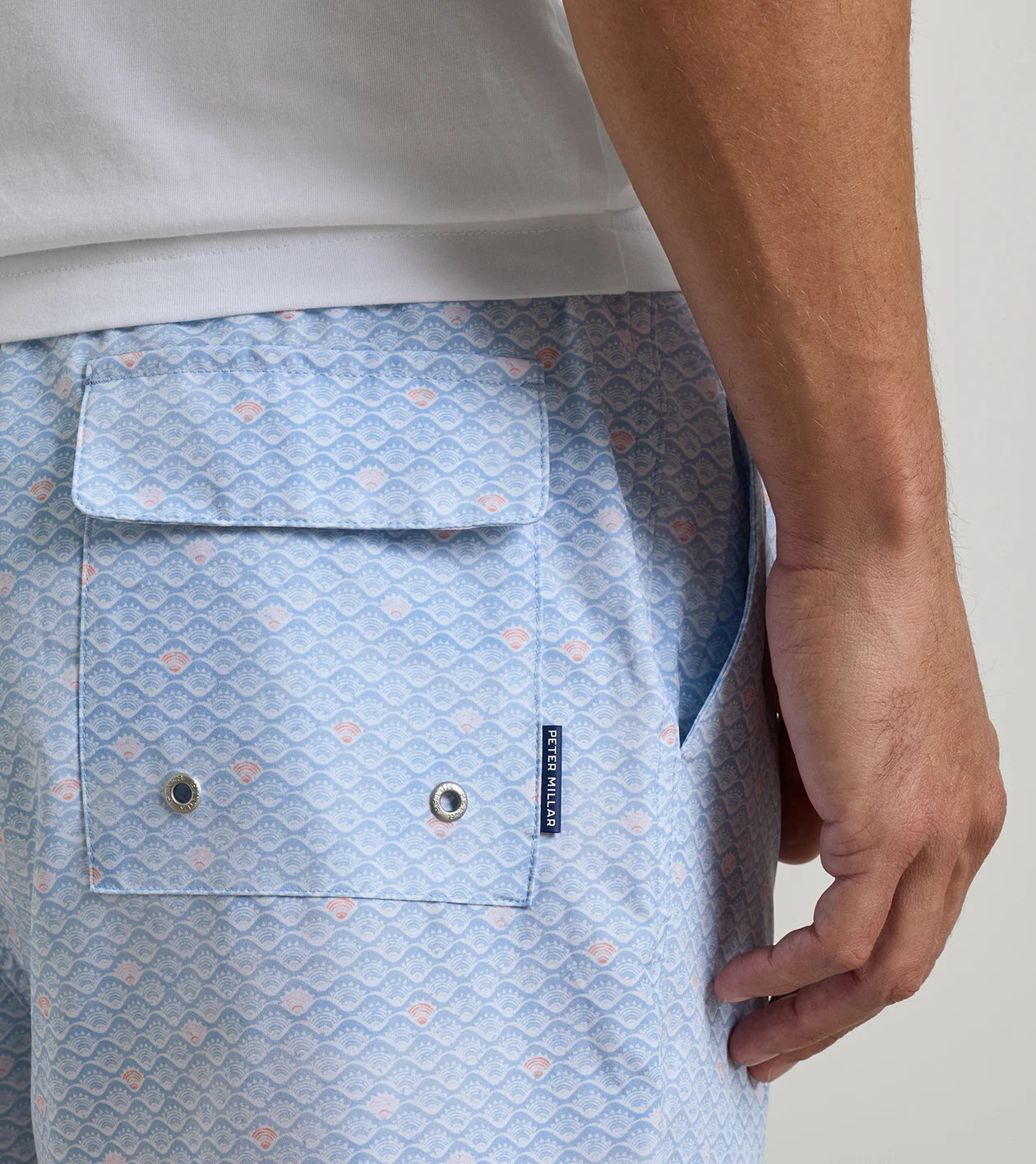 Wearing the Peter Millar Shoreline Rays Swim Trunk in a light blue patterned design, this person sports shorts with a back pocket, metal grommets, and a small navy label. Crafted from quick-drying fabric with an elastic drawstring waistband for comfort, the look is completed with a partially visible white shirt.