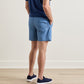 A man is seen standing indoors on a wooden floor, viewed from behind, wearing a blue shirt and Peter Millar Island Palms Swim Trunk in light blue—featuring a lightweight, quick-drying fabric with elastic drawstring waistband, mesh liner, and back pocket—paired with navy sneakers.