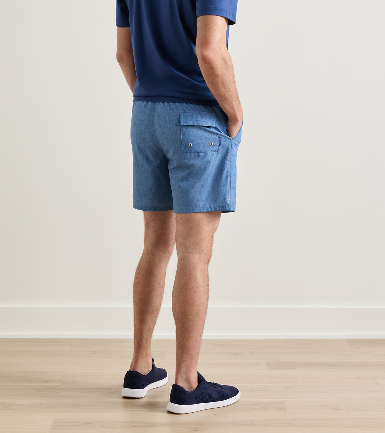A man is seen standing indoors on a wooden floor, viewed from behind, wearing a blue shirt and Peter Millar Island Palms Swim Trunk in light blue—featuring a lightweight, quick-drying fabric with elastic drawstring waistband, mesh liner, and back pocket—paired with navy sneakers.