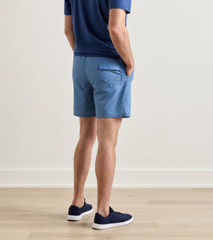 A man is seen standing indoors on a wooden floor, viewed from behind, wearing a blue shirt and Peter Millar Island Palms Swim Trunk in light blue—featuring a lightweight, quick-drying fabric with elastic drawstring waistband, mesh liner, and back pocket—paired with navy sneakers.