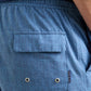 Close-up of the back pocket on the Peter Millar Island Palms Swim Trunk, featuring lightweight, quick-drying blue patterned shorts with a flap, two metal eyelets, a small “Peter Millar” brand tag, and an elastic drawstring waistband for enhanced comfort.