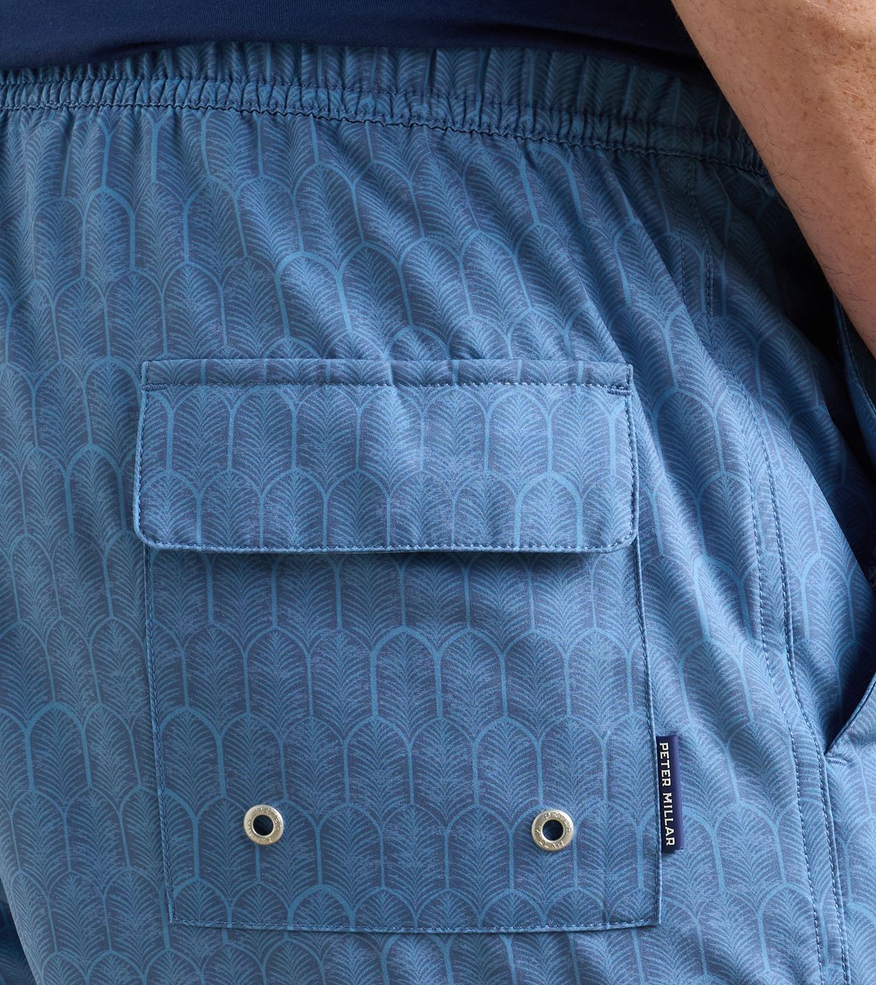 Close-up of the back pocket on the Peter Millar Island Palms Swim Trunk, featuring lightweight, quick-drying blue patterned shorts with a flap, two metal eyelets, a small “Peter Millar” brand tag, and an elastic drawstring waistband for enhanced comfort.