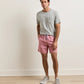 A man stands indoors in front of a plain beige wall on a light wood floor, wearing a grey t-shirt, grey sneakers, and Peter Millar Tails of Summer Swim Trunk featuring pink patterned fabric and an elastic drawstring waistband.